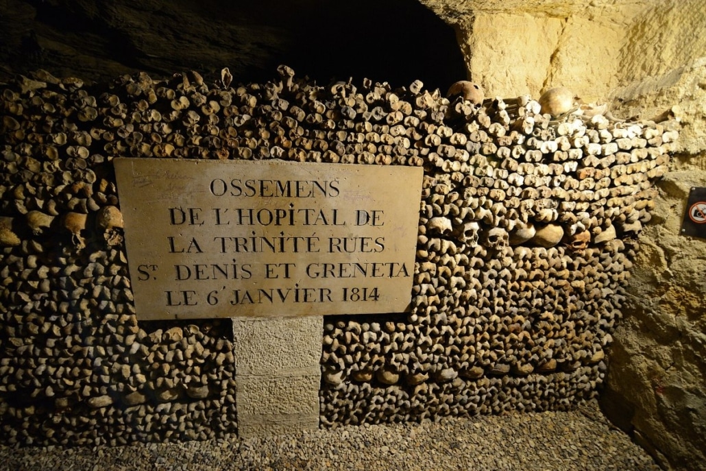 Een wand met menselijke botten en schedels in de Catacombes van Parijs, met een bordje dat de herkomst van de beenderen verme